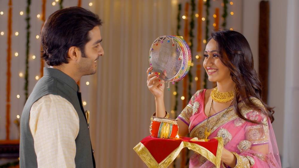 Young Indian couple happily performing Karwa Chauth traditions in traditional wear. Beautiful wife looking at the moon through a sieve and then to her good looking husband, following Karwa Chauth r...