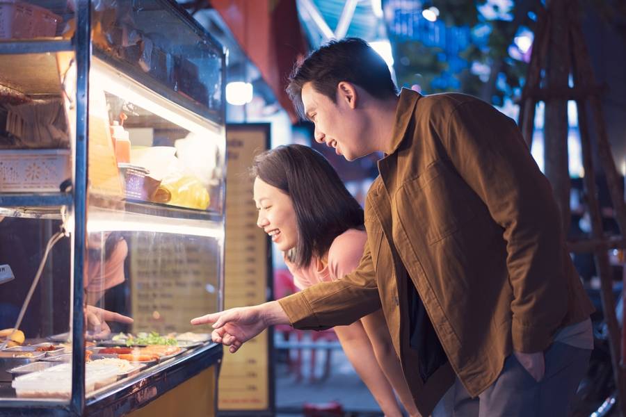 Two young individuals share laughter while selecting delicious street food from a vendor in Hanoi at night, surrounded by lively city lights and flavors.
