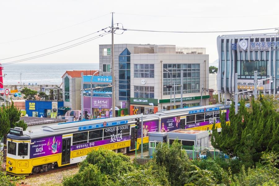 Green Haeundae Beach Train passing through Cheonsapo coastal area with cafés, shops, and ocean backdrop in Busan.