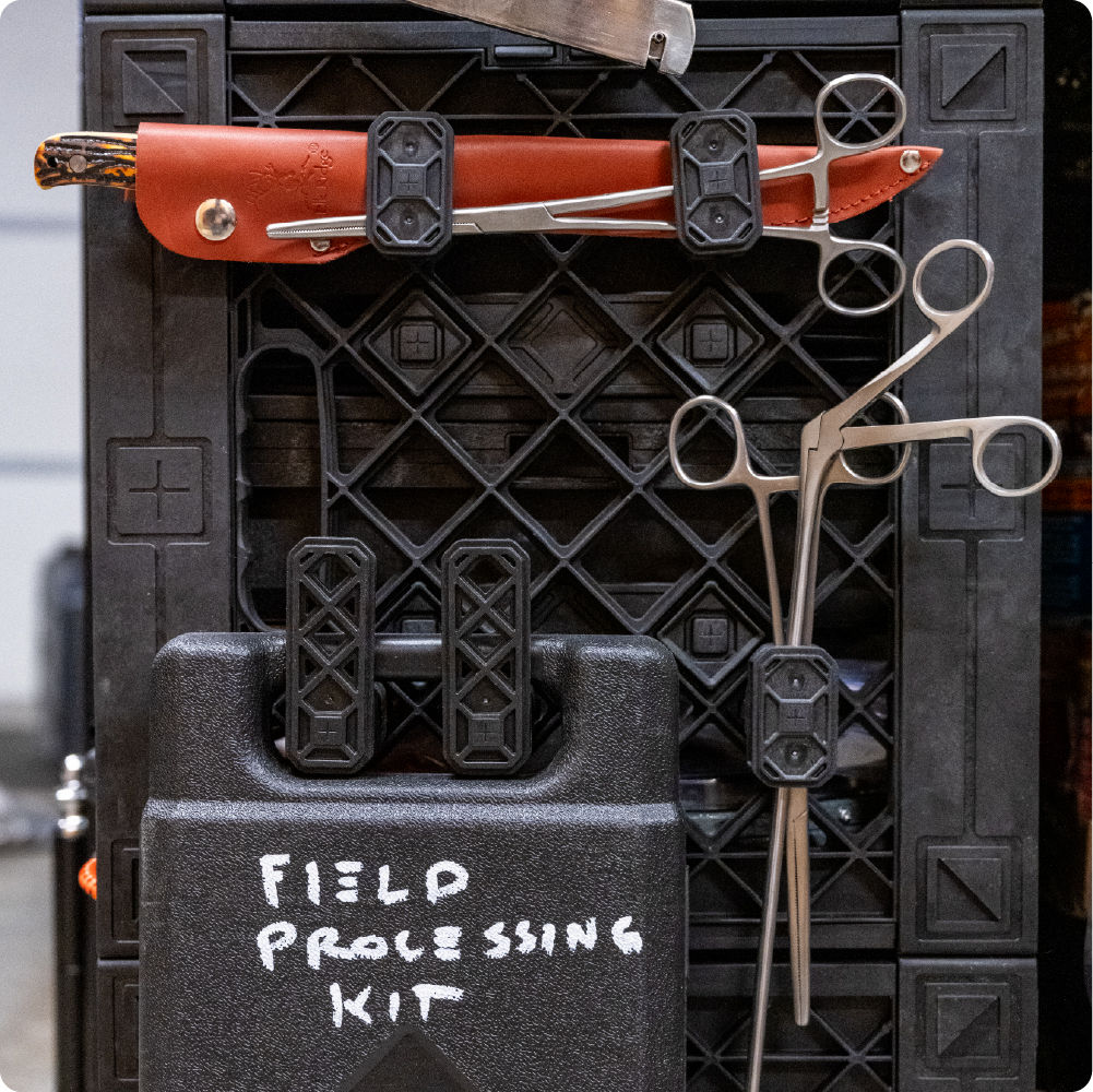 Close-up of medical and field-processing tools secured to the side of a black UbeCube crate with modular clips, including forceps, scissors, a knife sheath, and a labeled field processing kit.