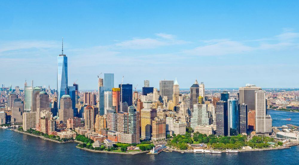 A breathtaking aerial view of New York City skyline, featuring iconic landmarks such as the Empire State Building, One World Trade Center and the Hudson River. The city is illuminated with a golden glow, creating a stunning visual display.