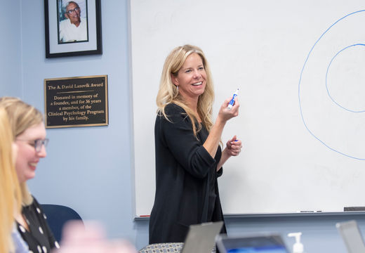 A professor smiling while teaching at a whiteboard in a classroom.