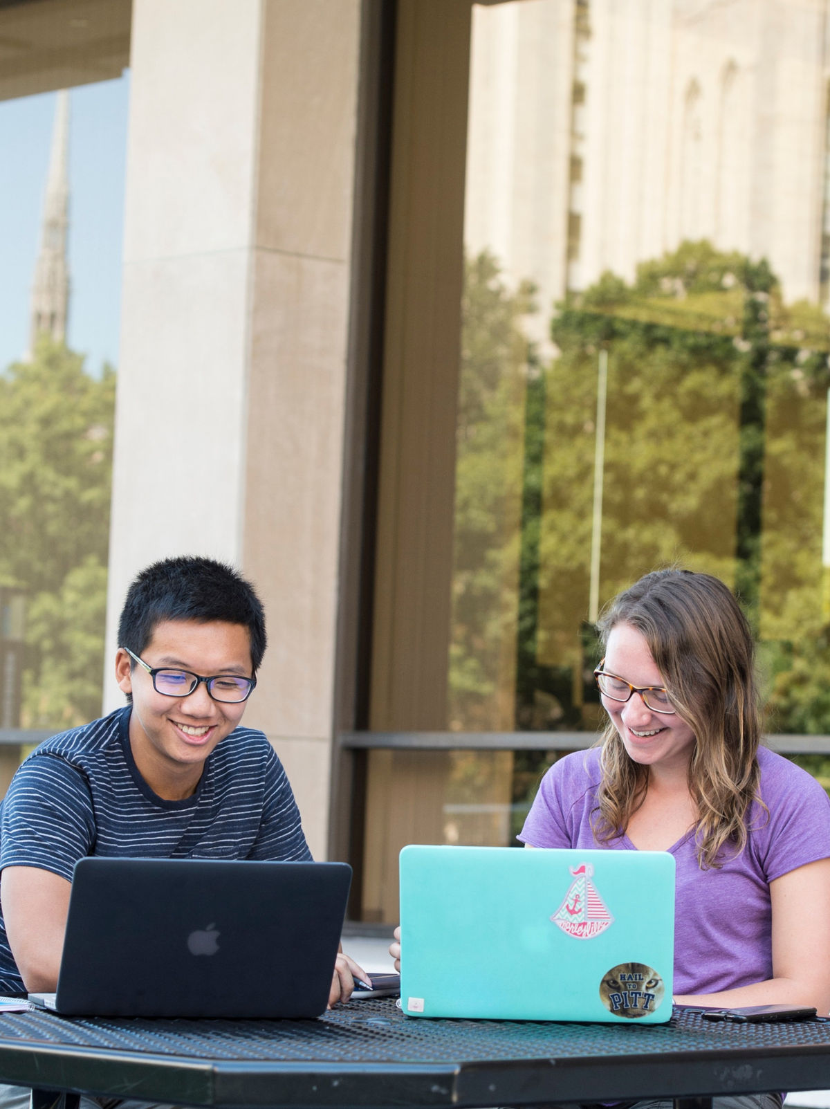 Two students studying on laptops at an outdoor table on campus.