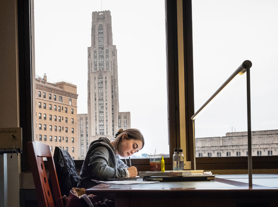 A student studying at a library desk with the Cathedral of Learning visible through the window.