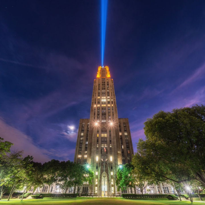 The University of Pittsburgh's Cathedral of Learning illuminated at night with blue and gold light beams.
