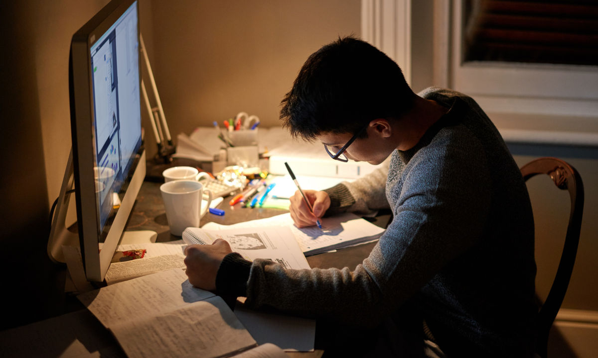 A student studying at a desk late at night, writing in a notebook with a computer nearby.