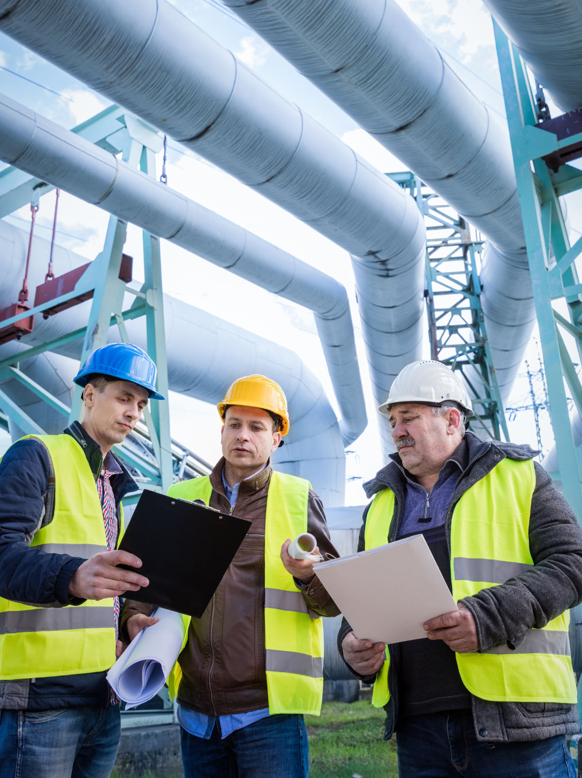 Three people in hard hats and safety vests reviewing documents at an industrial facility.