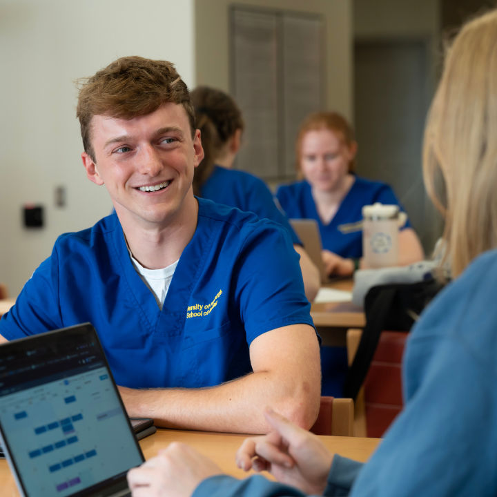 Nursing students in scrubs talking and working on laptops in a common area.