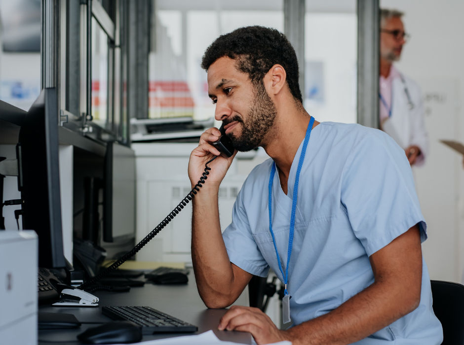 A healthcare professional in scrubs talking on the phone at a workstation.