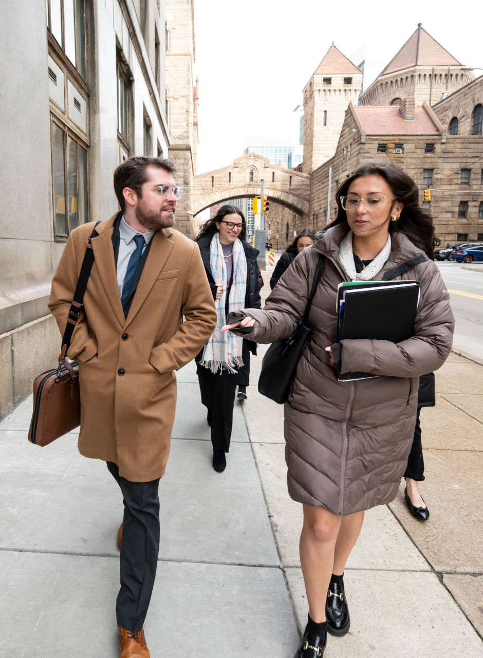 Students walking along a city sidewalk near university buildings, carrying bags and notebooks.