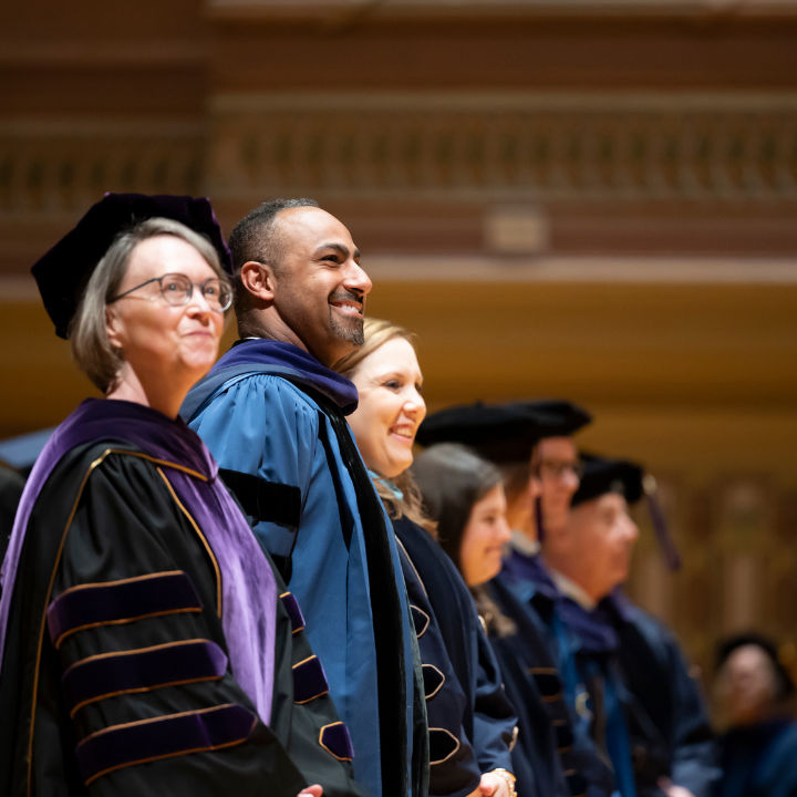 A row of faculty in academic regalia smiling during a graduation ceremony.