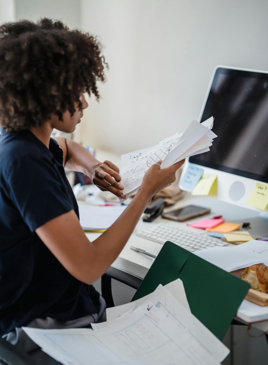 A person reviewing handwritten notes at a desk with a computer and scattered papers and sticky notes.