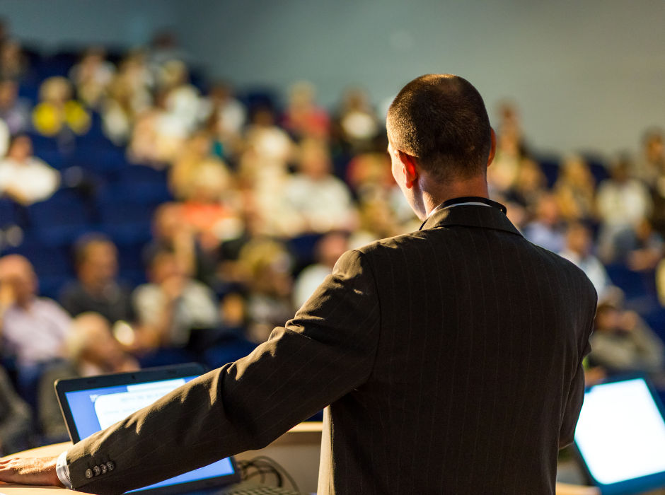 A speaker at a podium addressing a large audience in a lecture hall, viewed from behind.