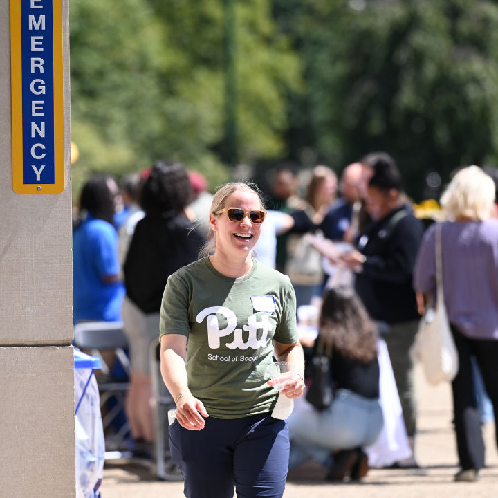 A smiling person wearing a Pitt School of Social Work t-shirt at an outdoor campus event.