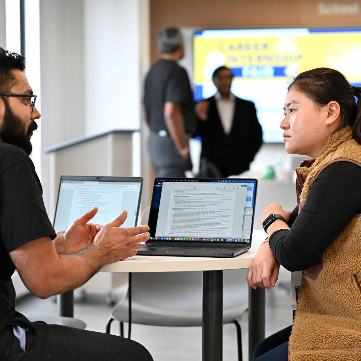 Two people in conversation at a table with laptops.