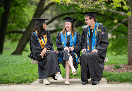 Three graduates in caps and gowns chatting on a campus bench.