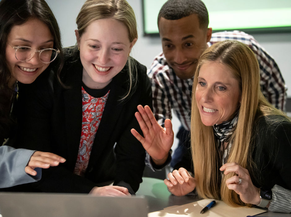 Four people gathered around a laptop, smiling and engaged in what they're viewing.