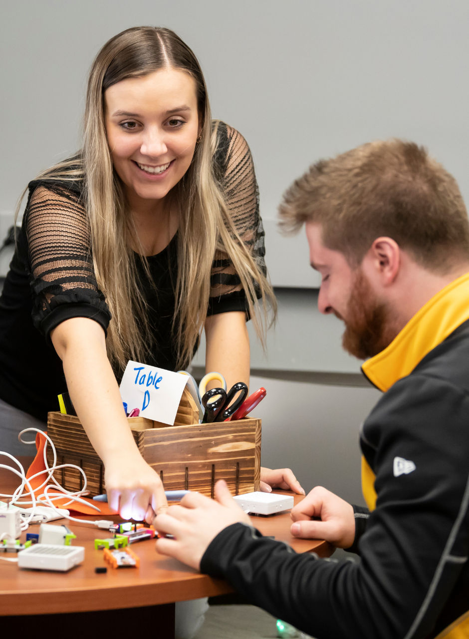 Two people working together at a table with small electronic components and craft supplies.