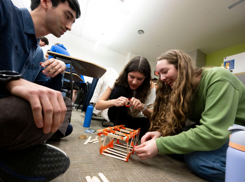 Three students sitting on the floor collaborating on building a small structure with craft sticks and tape.