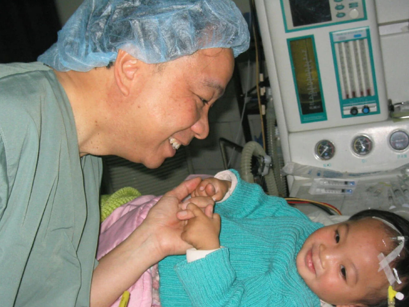 Doctor wearing surgical cap smiling and holding the hand of a baby lying in a hospital bed with medical equipment in the background