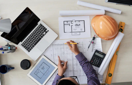 A top down view of a construction worker viewing blueprints beside a laptop.