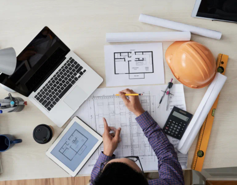 A top down view of a construction worker viewing blueprints beside a laptop.