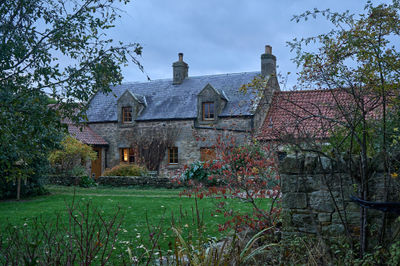 A charming stone house with a rustic appearance sits amidst a well-maintained lawn and tall trees, creating a serene and inviting atmosphere for a peaceful retreat.