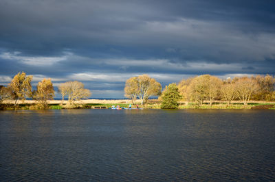 Musselburgh Lagoons. A serene scene: a boat sails on calm waters surrounded by trees in a vast field. Cloudy sky adds drama. People enjoy the peaceful environment.