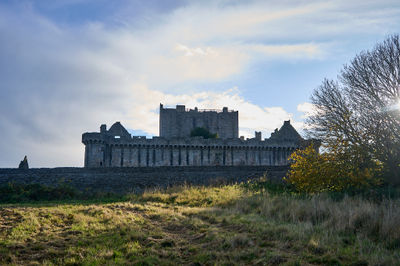 Craigmillar Castle. Stone castle surrounded by a wall and green field. Tree grows on wall, adding nature. Building with a fence nearby. Leafless tree contrasts.