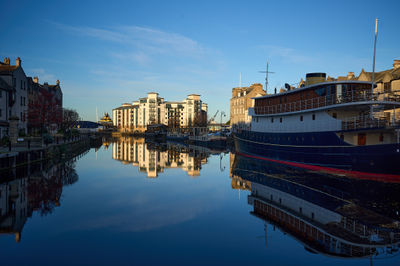 Water Of Leith Shore. A photo of a boat gliding on calm water, with detailed craftsmanship and a group of buildings reflecting sunlight.