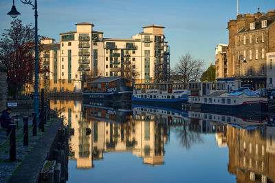 Water Of Leith Shore. A peaceful waterfront scene with a building and boats. One boat stands out with multiple windows, suggesting a larger size.