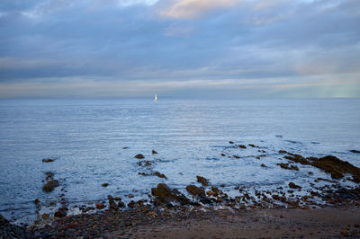 A serene coastal scene with a rocky beach, a sailboat gliding across tranquil waters, rugged terrain, and a serene sky adorned with fluffy clouds.