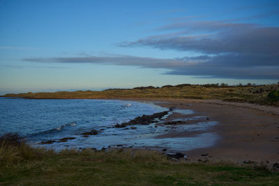 Gullane, East Lothian, Scotland. A serene beach scene: rocky shore, calm waters, blue sky, sandy stretches, scattered rocks, grassy field.