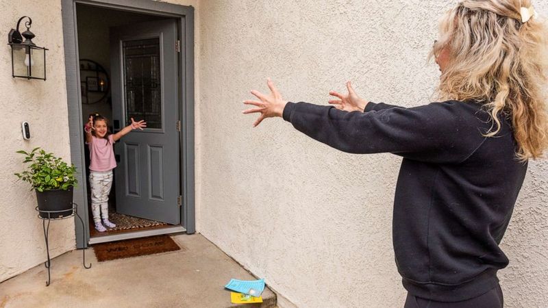 Social distancing photos - kindergartener student at the door and her teacher exchanging hugs from a distance