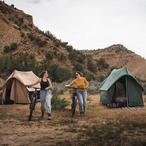 Two women enjoying mountain biking near camping tents in a scenic outdoor setting