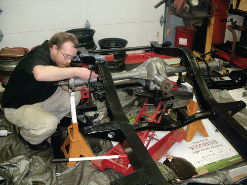 With the axle supported on a Harbor Freight trans jack, Jesse bolts the right-hand check strap to the bottom of the frame. The axle was painted with gray ceramic engine paint.