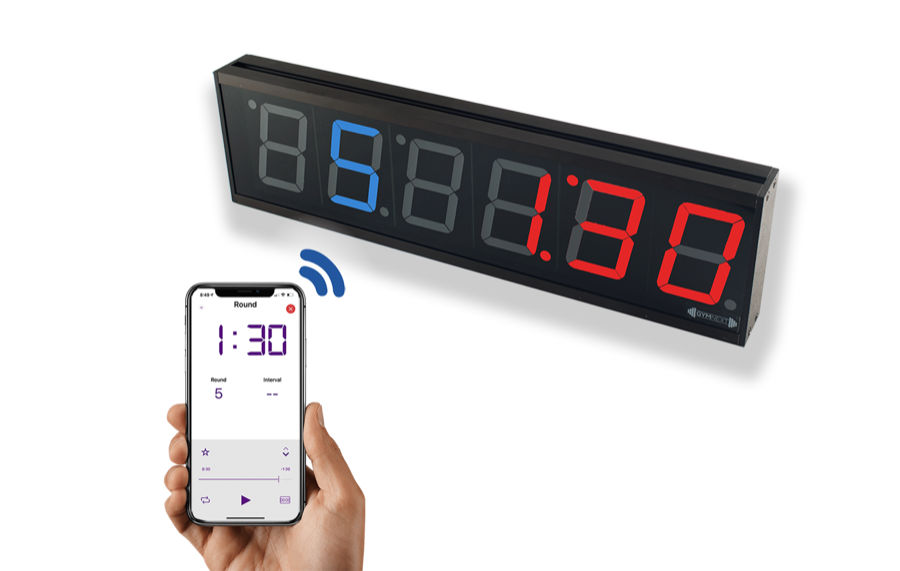 GymNext Flex Timer, a long black six-digit LED wall clock showing 1:30 in red and 5 in blue, paired over Bluetooth with a phone app held in a hand