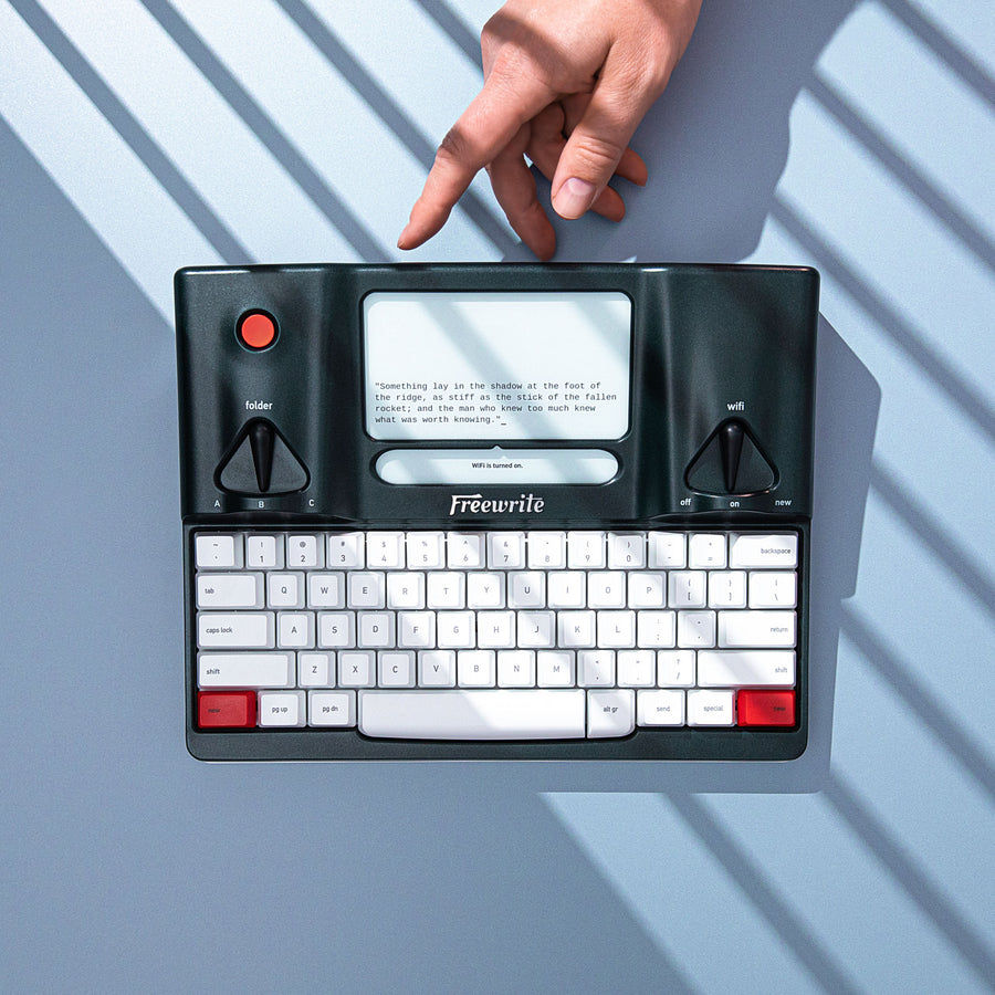 Freewrite Smart Typewriter in dark green photographed from above in hard shadow, with two chrome dials flanking a frontlit e-ink screen above white mechanical keys