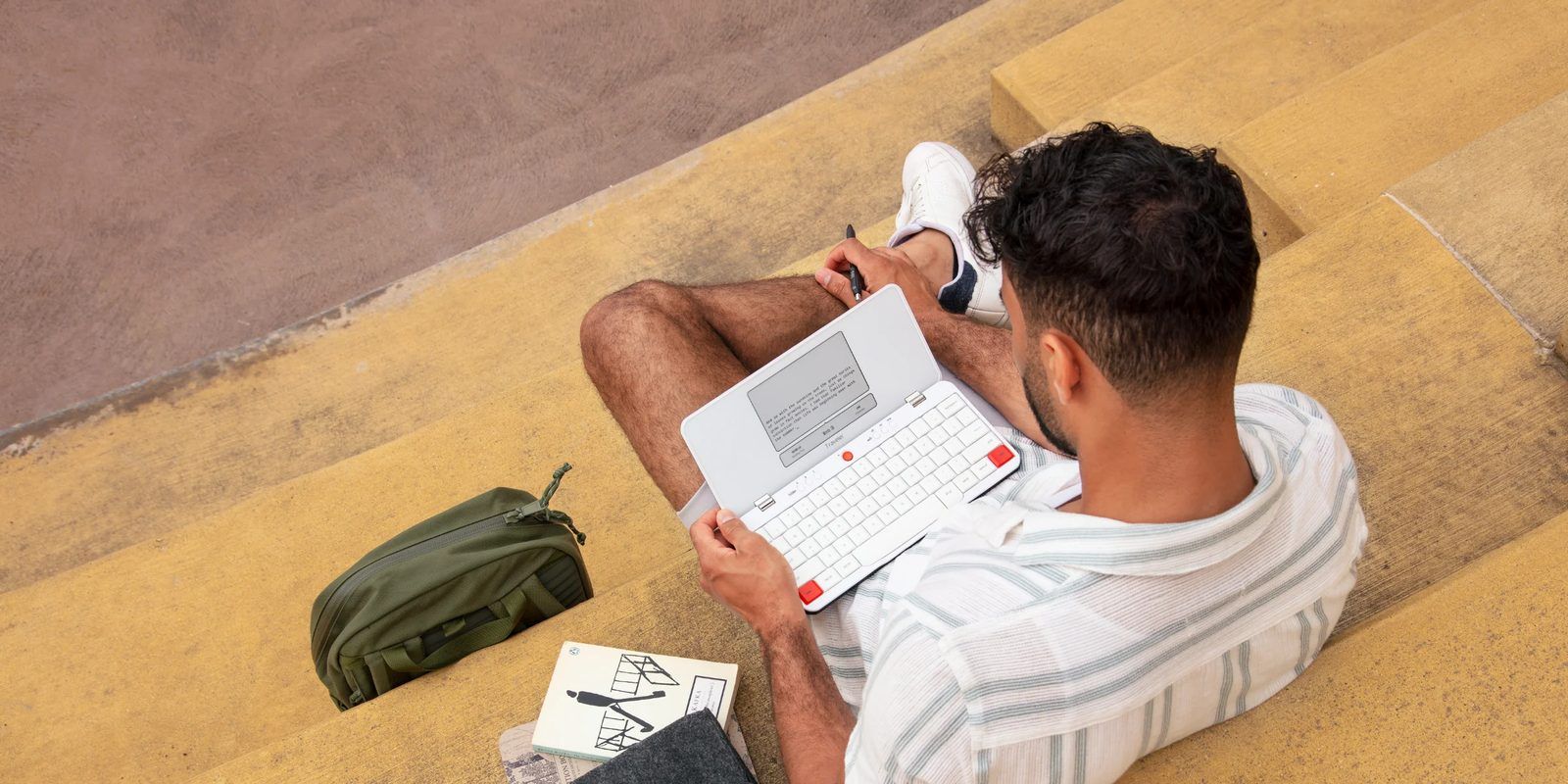 Person sitting on concrete steps typing on a white Freewrite Traveler, an e-ink clamshell writing device, with a book and bag beside them