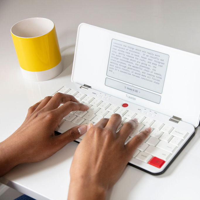 Hands typing on a white Freewrite Traveler open on a desk, its angled e-ink screen showing a paragraph of drafted text