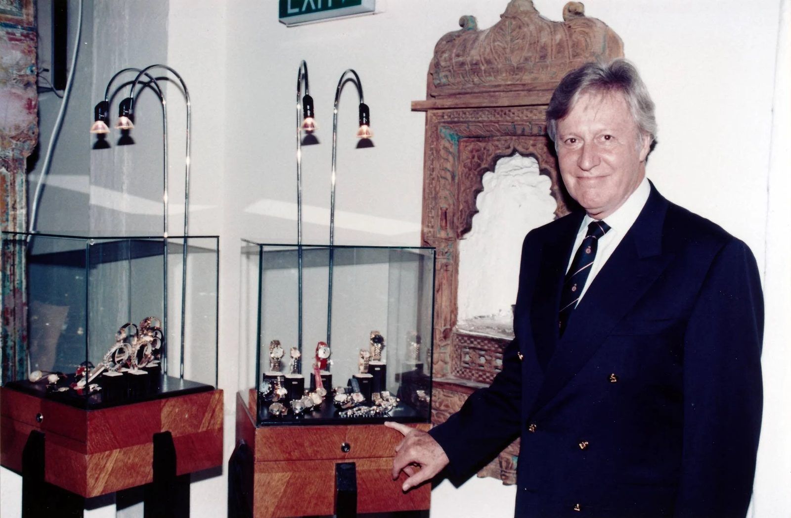 Gérald Genta, grey-haired, in a navy double-breasted blazer and striped tie, smiling and pointing at a lit glass display case of his watches in a shop