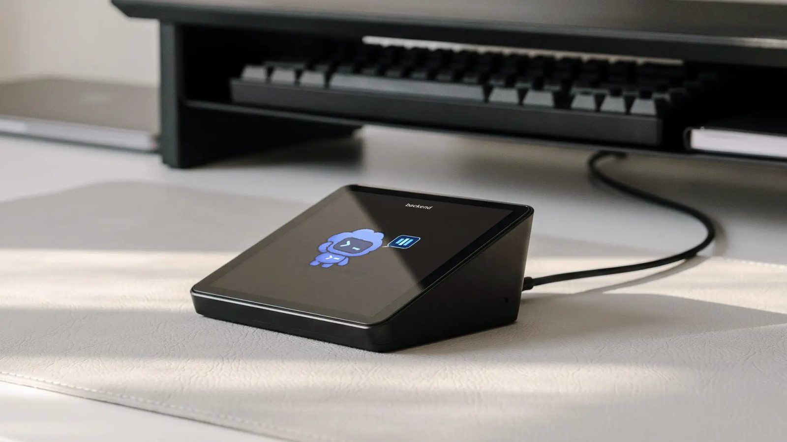 Black Harness Pro on a white desk mat in front of a keyboard, its tilted square screen showing a blue robot pet for the agent named backend