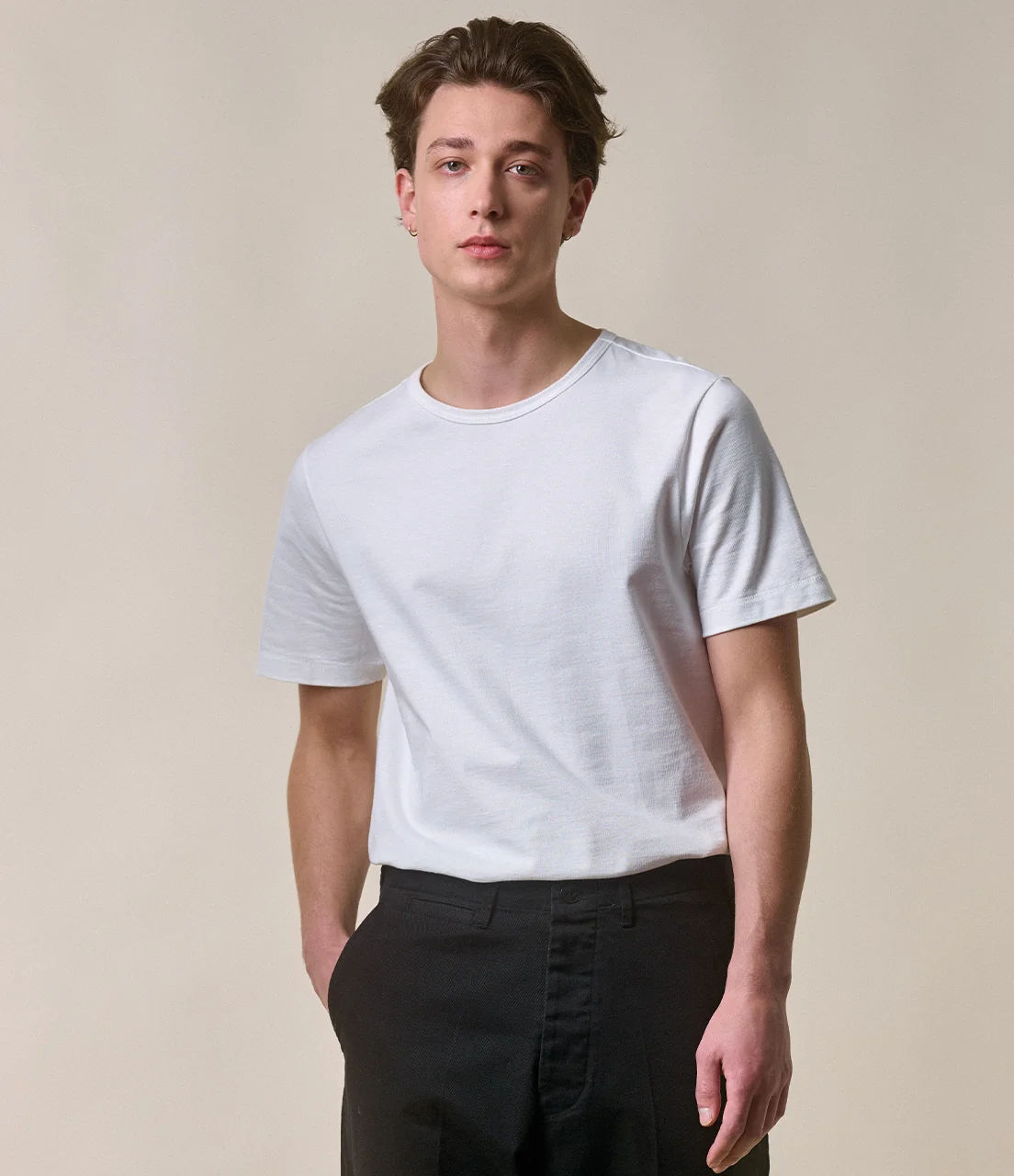 Man wearing a plain white lighter-weight Merz crew neck tee with black trousers against a beige studio background