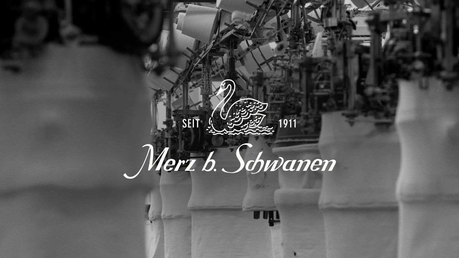 Black and white photo of a hall of vintage loopwheel knitting machines with tubes of white jersey hanging beneath them, overlaid with the Merz b. Schwanen swan logo and SEIT 1911