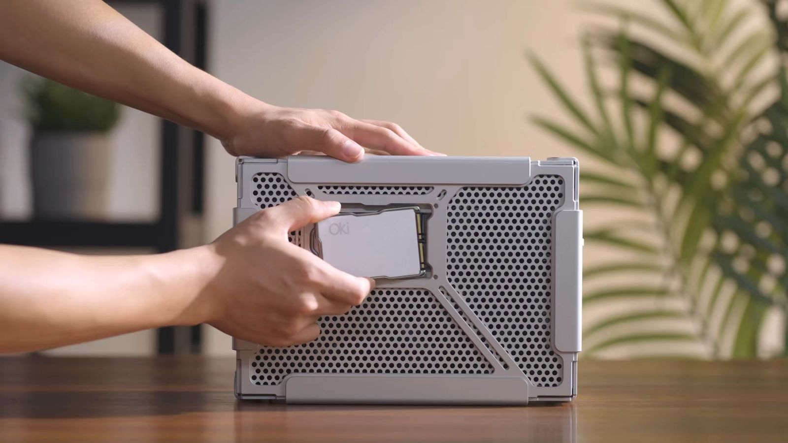 2 hands on top of an Oki Home sitting on a wooden table, one pushing a silver Memchip card into the slot window on its perforated aluminium front, with a plant in the background