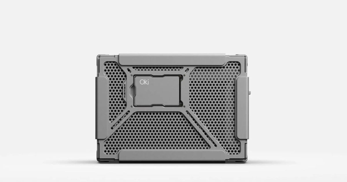Oki Home seen from the front on a white background: a wide bead-blasted aluminium box with a perforated hexagon-mesh face, a raised X-shaped frame across it and a small slot window holding a card marked Oki