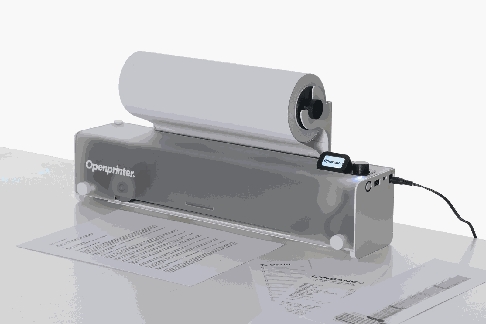 OpenPrinter on a desk: a slim grey body with a paper roll mounted on top, a small screen and control knob, and printed pages in front