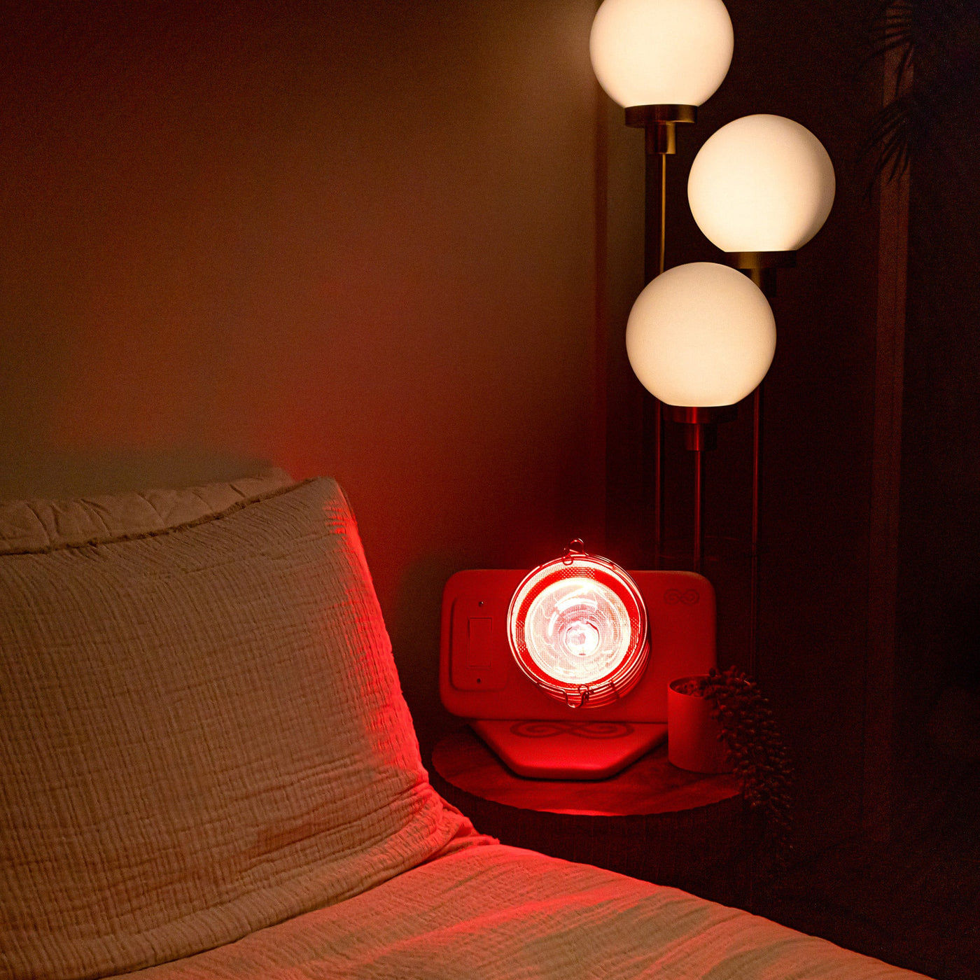 The Glow on a bedside table in a dark bedroom, casting red light across the pillows next to a floor lamp with 3 white globes