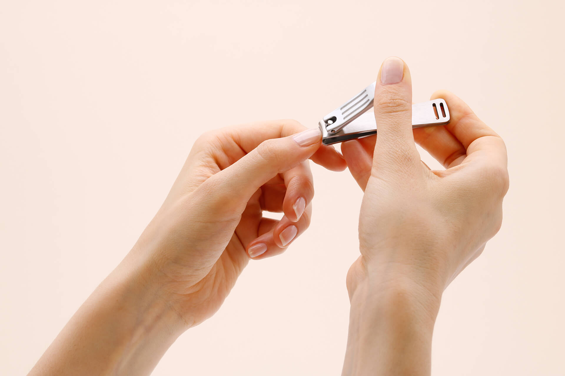 A pair of hands against a pale pink background, one hand trimming a fingernail of the other with a small silver Seki Edge clipper