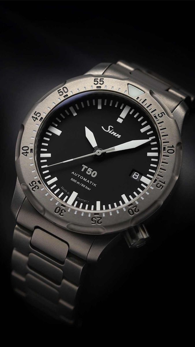 Close-up of the Sinn T50 on its titanium bracelet against a black background: scalloped bead-blasted bezel with a luminous triangle, black dial with large white sword hands, date at 4 o'clock and 500 m / 50 bar on the dial
